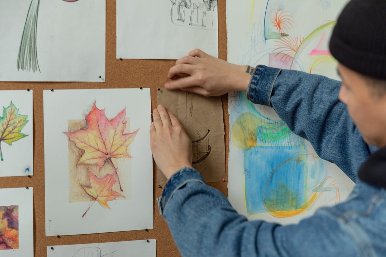 A person pins sketches on a corkboard, showcasing autumn leaf drawings in an artistic setup.